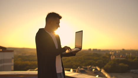 The-camera-follows-a-man-walking-across-a-rooftop-at-sunset-with-a-laptop-and-a-bottle.-The-freelancer-opens-the-laptop-and-types-on-the-keyboard.-Slow-motion-hacker-walks-on-the-roof-with-a-laptop.
